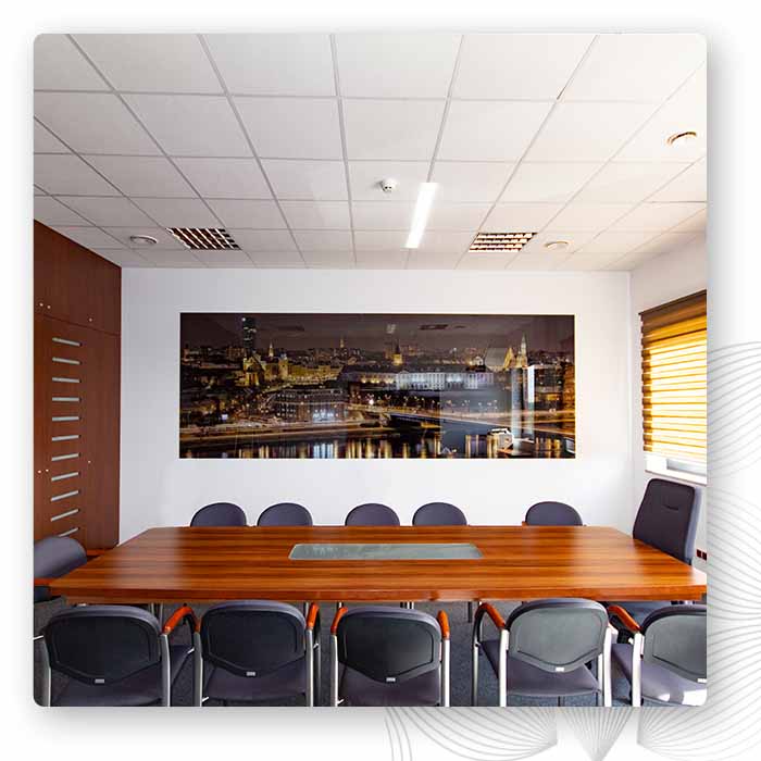 Sala konferencyjna z dużym drewnianym stołem i czarnymi fotelami. Na białej, centralnej ścianie wisi szeroki szklany panel z nocną panoramą miasta."